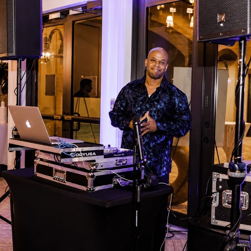 A DJ stands behind a table with audio equipment and a laptop, smiling at the camera in an indoor event space.