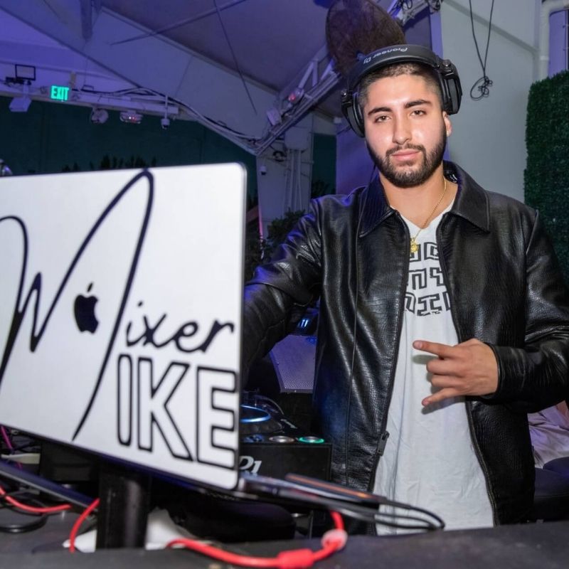 A DJ wearing headphones and a black jacket stands behind a laptop with "Mixer Mike" written on it, performing at an indoor venue.