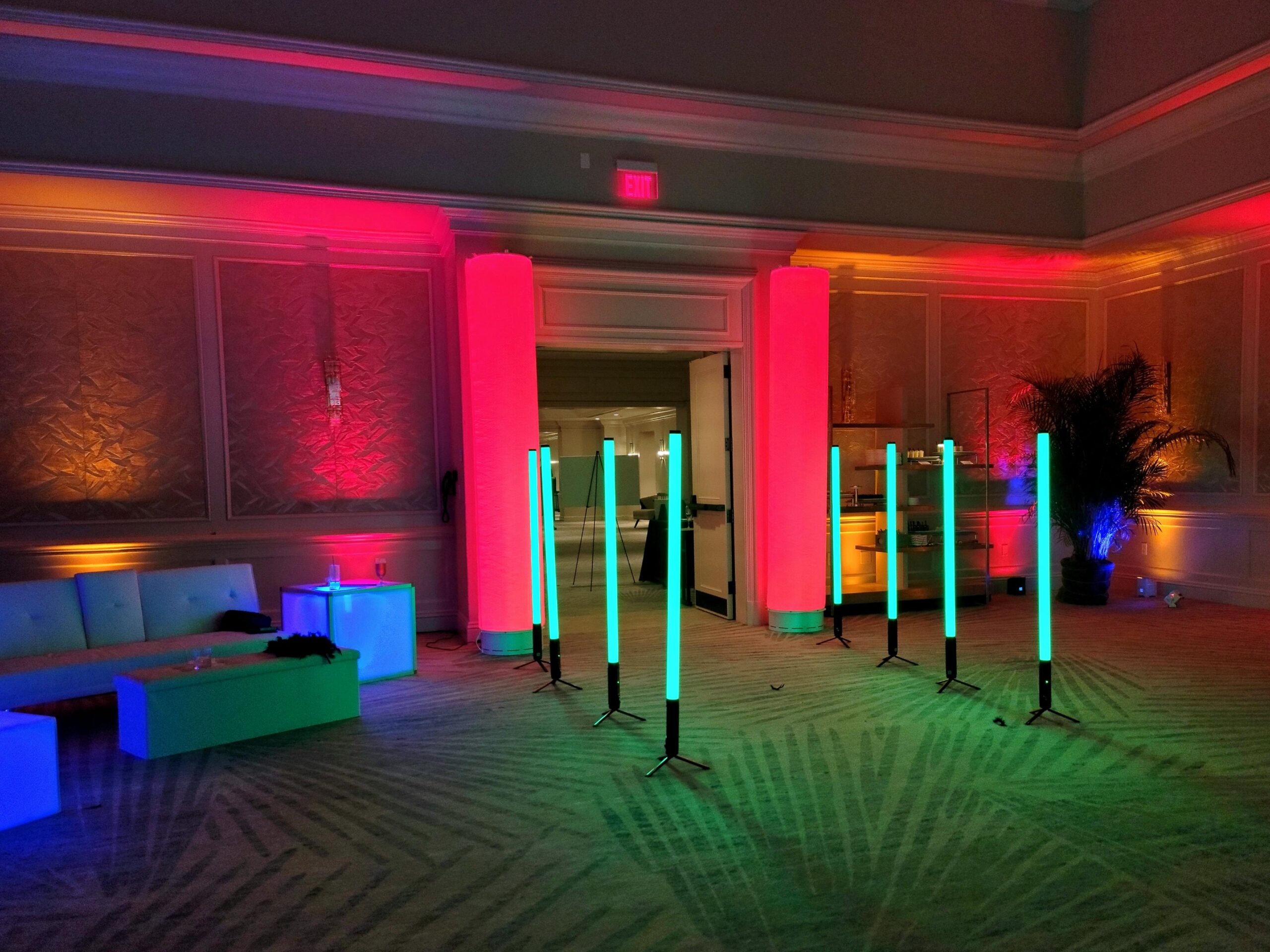 A dimly lit room with colorful LED pillars and tube lights, lounge seating, a palm plant, and decorative wall panels—perfect for stylish gatherings, with options for events add ons to elevate your experience.