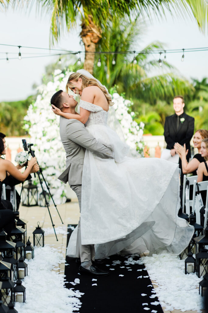 A groom lifts and kisses the bride at an outdoor wedding ceremony, surrounded by guests, white flower petals, palm trees, and the joyful energy of professional wedding DJ services filling the air with music.