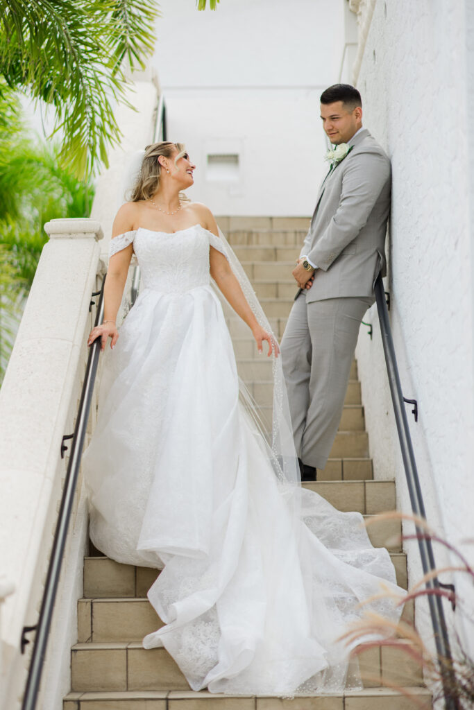 A bride in a white gown stands on stairs, smiling at a groom in a gray suit, who is leaning against the railing and looking at her as soft music from the wedding DJ services fills the air.