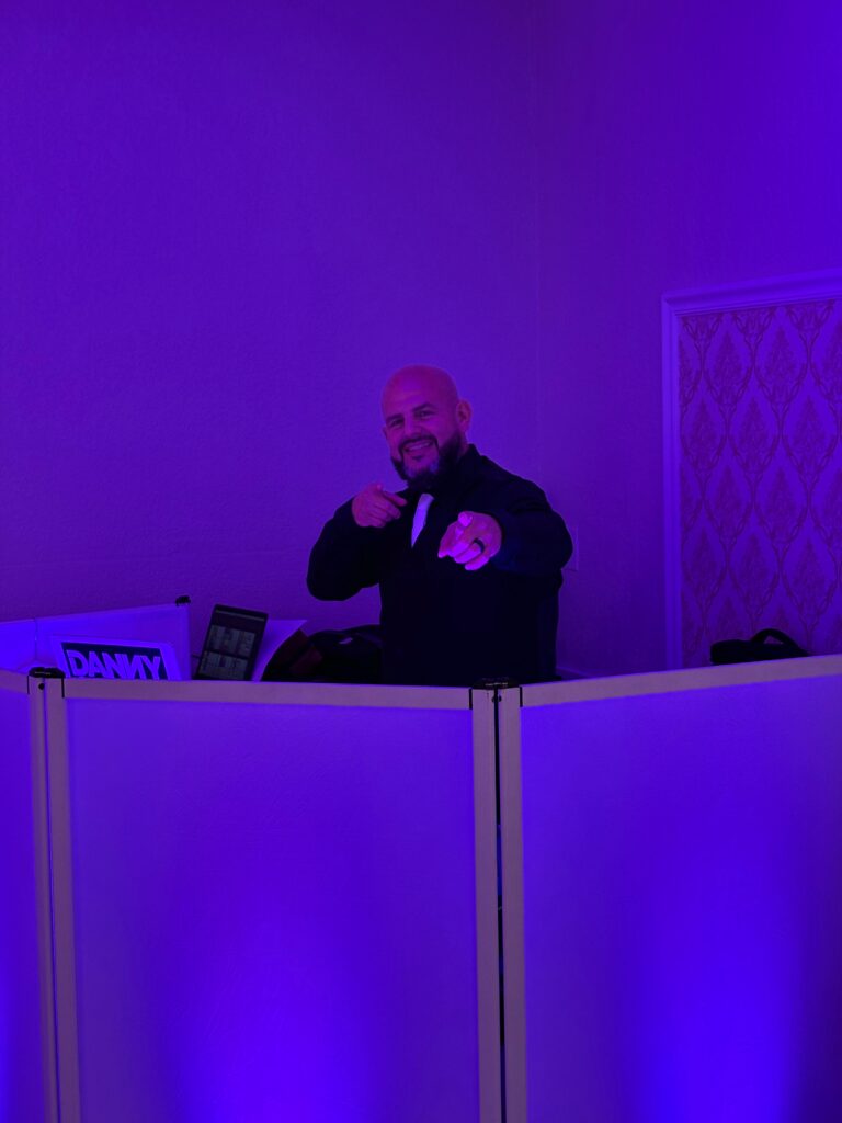 A man in a dark suit stands behind a DJ booth with purple lighting, smiling and pointing toward the camera in what appears to be a vibrant gallery setting.