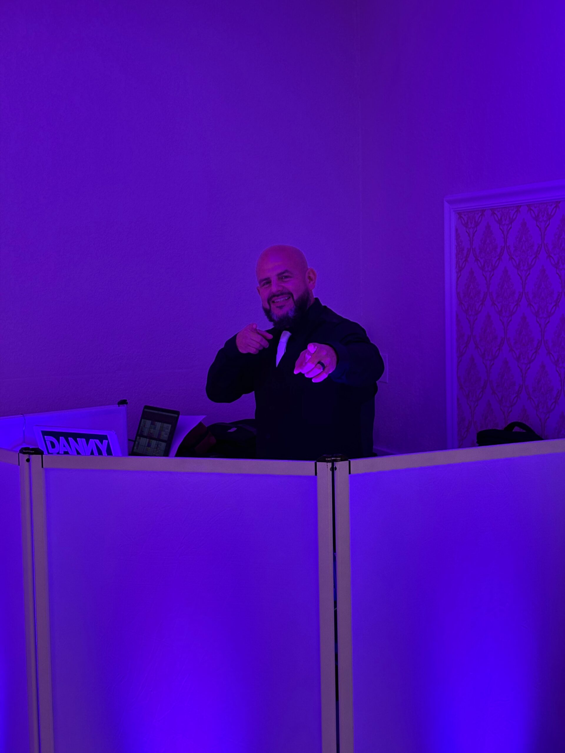 A man in a dark suit stands behind a DJ booth with purple lighting, smiling and pointing toward the camera in what appears to be a vibrant gallery setting.