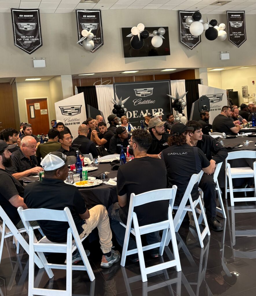 A group of people seated at round tables in a decorated room with Cadillac banners, attending a lively corporate event with food and drinks—perfectly complemented by the beats of a professional corporate events DJ.