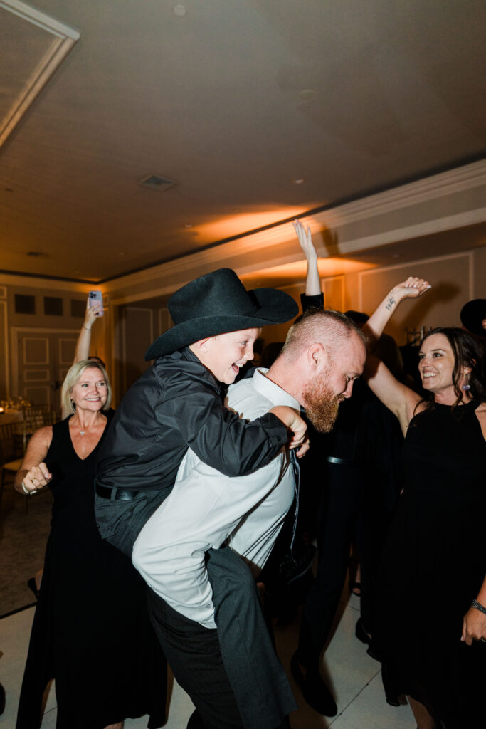 A man with a beard gives a boy in a cowboy hat a piggyback ride on a dance floor while people around them smile and raise their arms, enjoying the lively atmosphere created by the wedding DJ services.