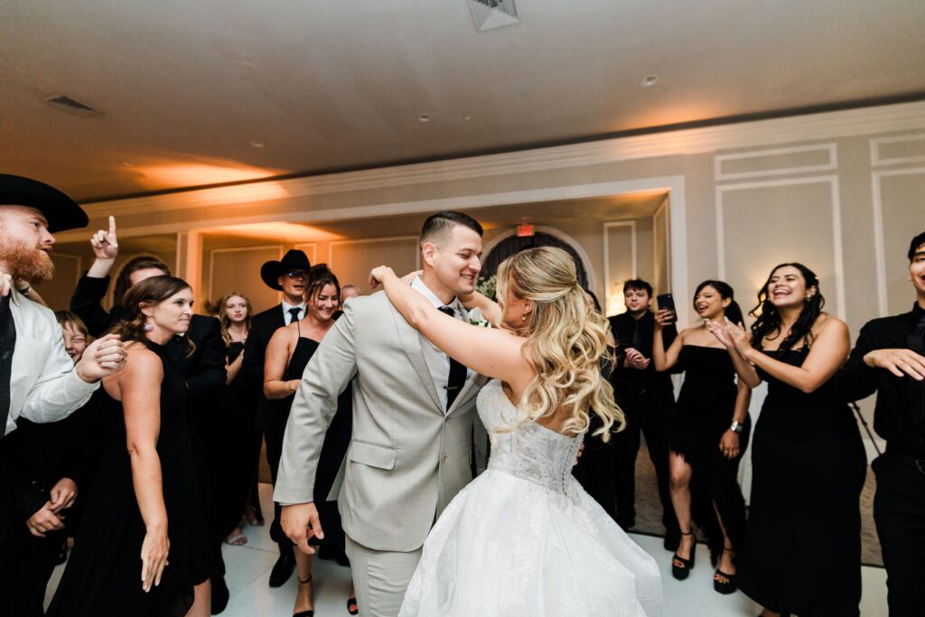 A bride and groom dance together, surrounded by guests in black attire, in a well-lit reception hall enhanced by lively wedding DJ services.