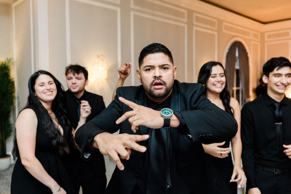 A man in a suit makes expressive hand gestures at a formal event, possibly discussing wedding DJ services, surrounded by four people dressed in black, all smiling and standing indoors.