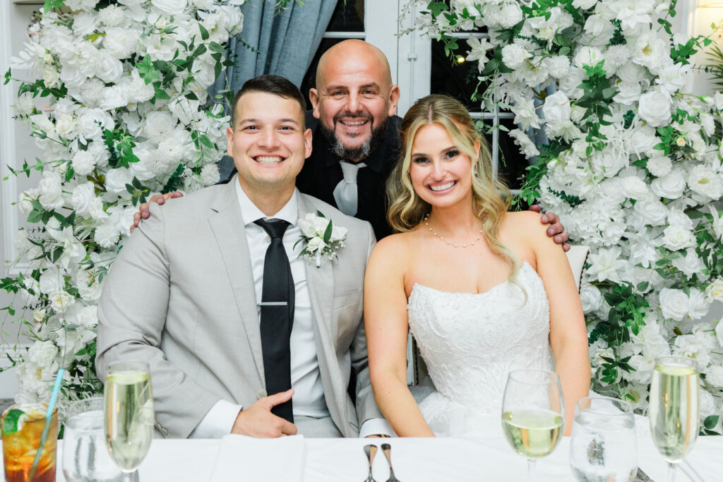 Three people, including a bride in a white dress and a groom in a light suit, sit at a table decorated with white flowers, smiling at the camera, as the joyful atmosphere is enhanced by professional wedding DJ services in the background.