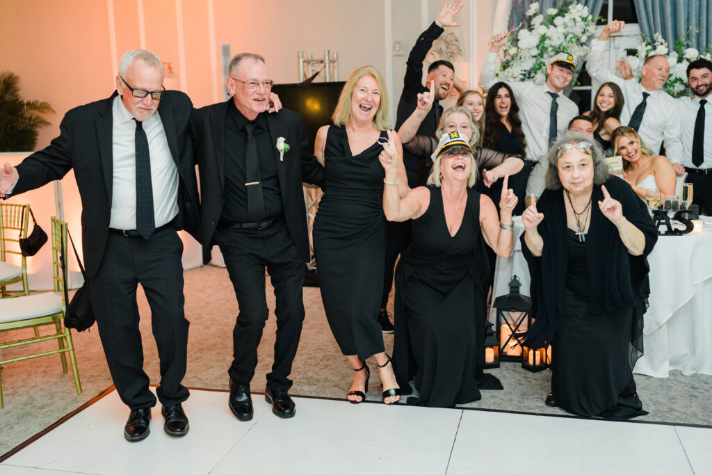 A group of people dressed in black celebrate together on a dance floor, smiling and posing with raised arms at an indoor event enhanced by exceptional wedding DJ services.
