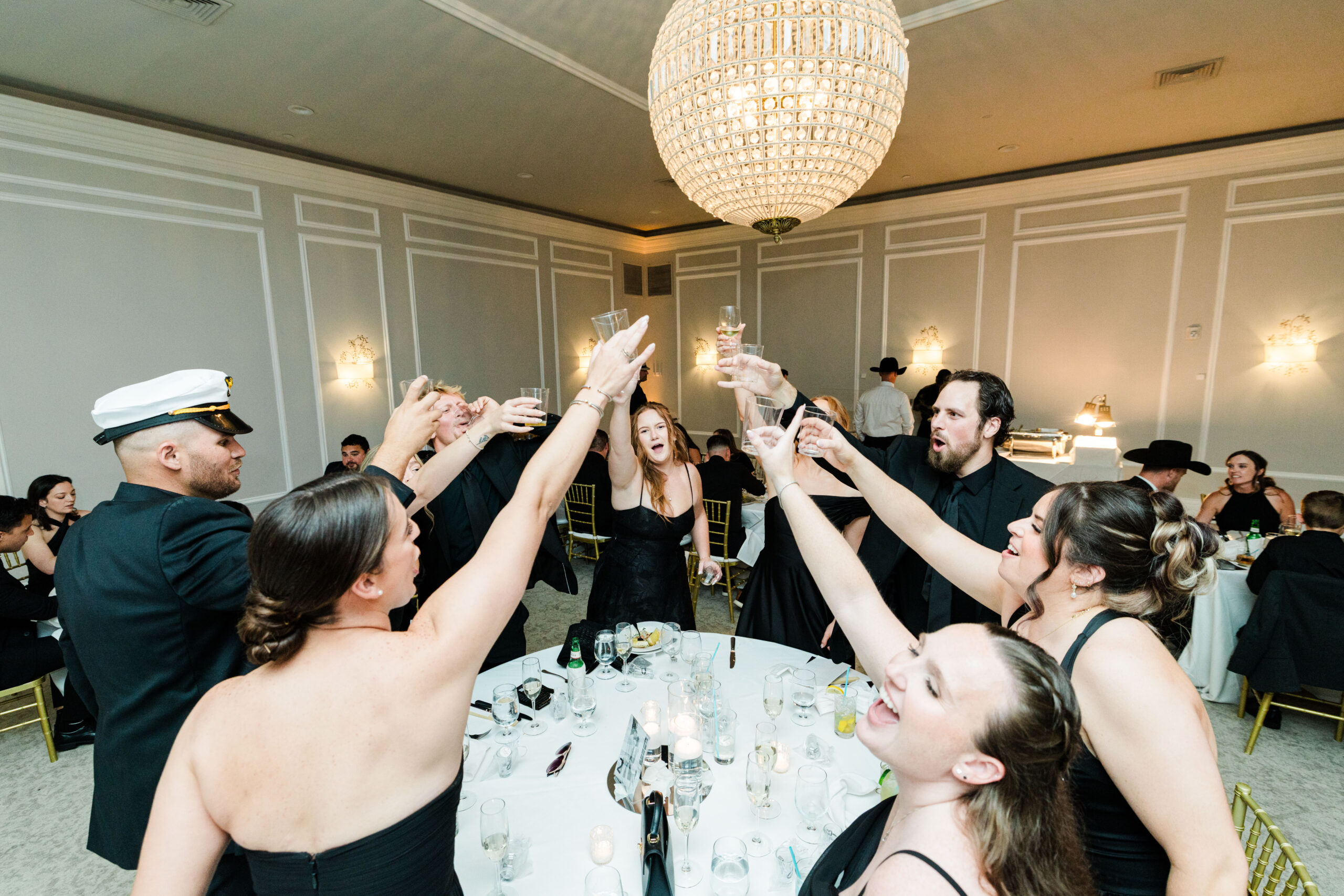 A group of people in formal attire stand around a table raising glasses in a toast at an indoor event, celebrating joyfully with music curated by professional wedding DJ services.