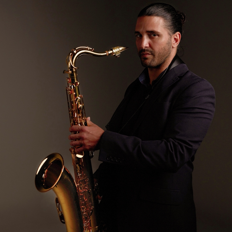 Man in a dark suit holds a brass saxophone against a plain brown background, looking at the camera with a neutral expression, as if inviting you to wonder about his story.