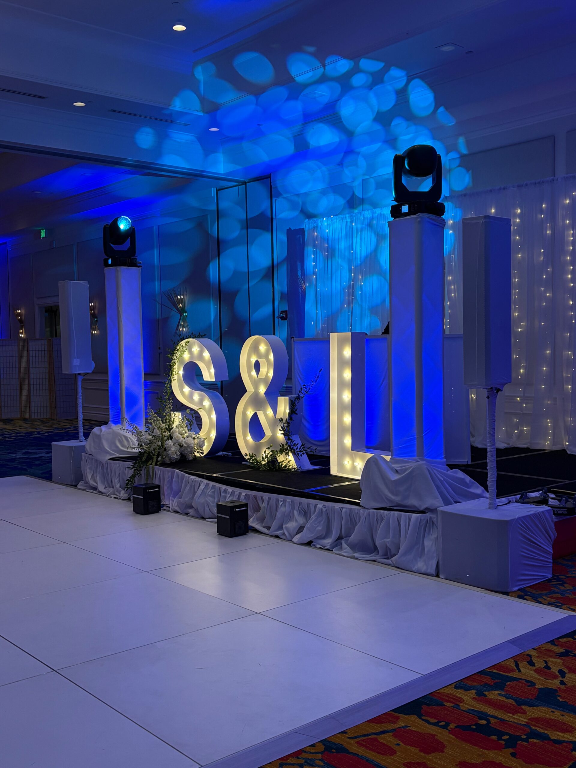 Large illuminated letters “S & L” on a decorated stage with blue lighting, surrounded by white drapes and speakers, set up as stylish events add ons for a celebration or special occasion in a ballroom.