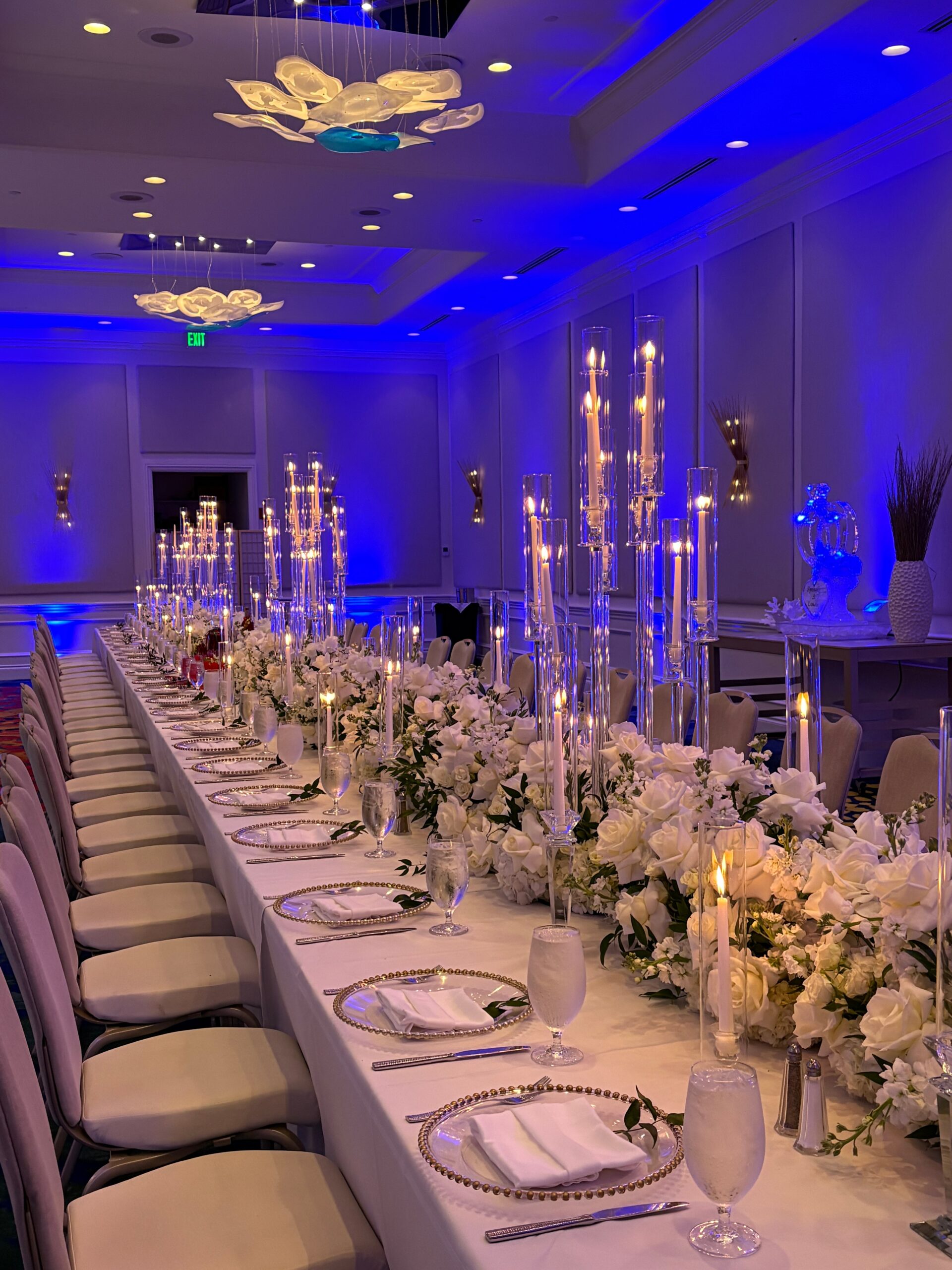 A long banquet table with white floral arrangements, tall candles, and place settings is set in an elegant gallery room with blue lighting and modern chandeliers.