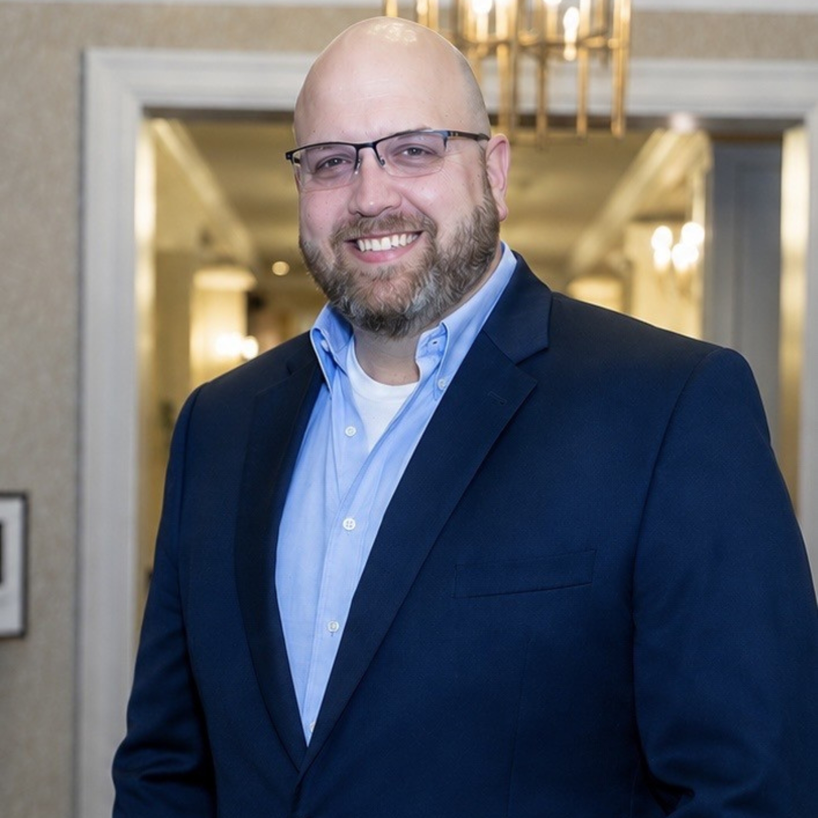 About: A man with glasses and a beard, wearing a navy blazer over a light blue shirt, stands indoors and smiles at the camera.